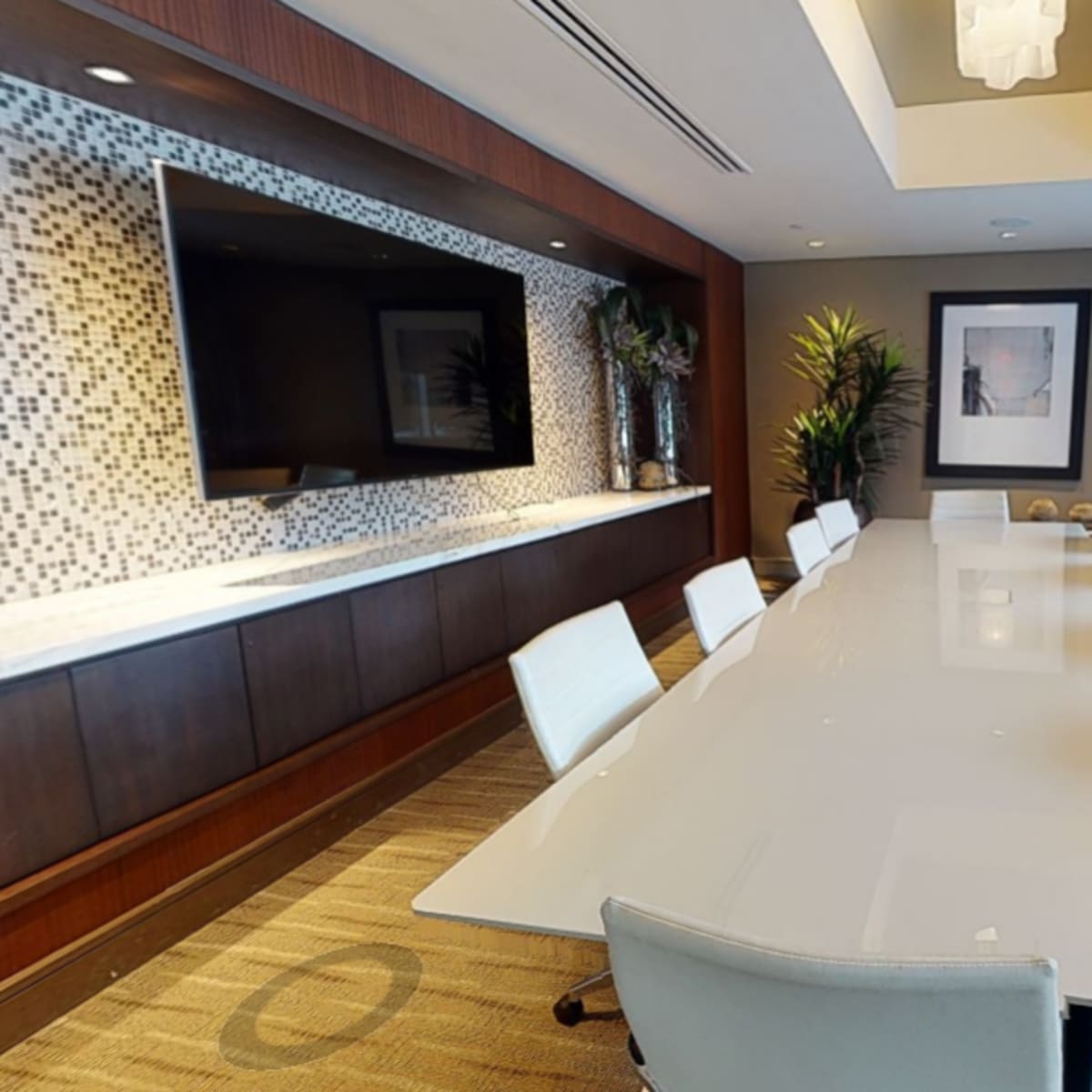 A modern conference room features a long white table surrounded by sleek chairs, with large windows providing natural light and a television mounted on the wall.