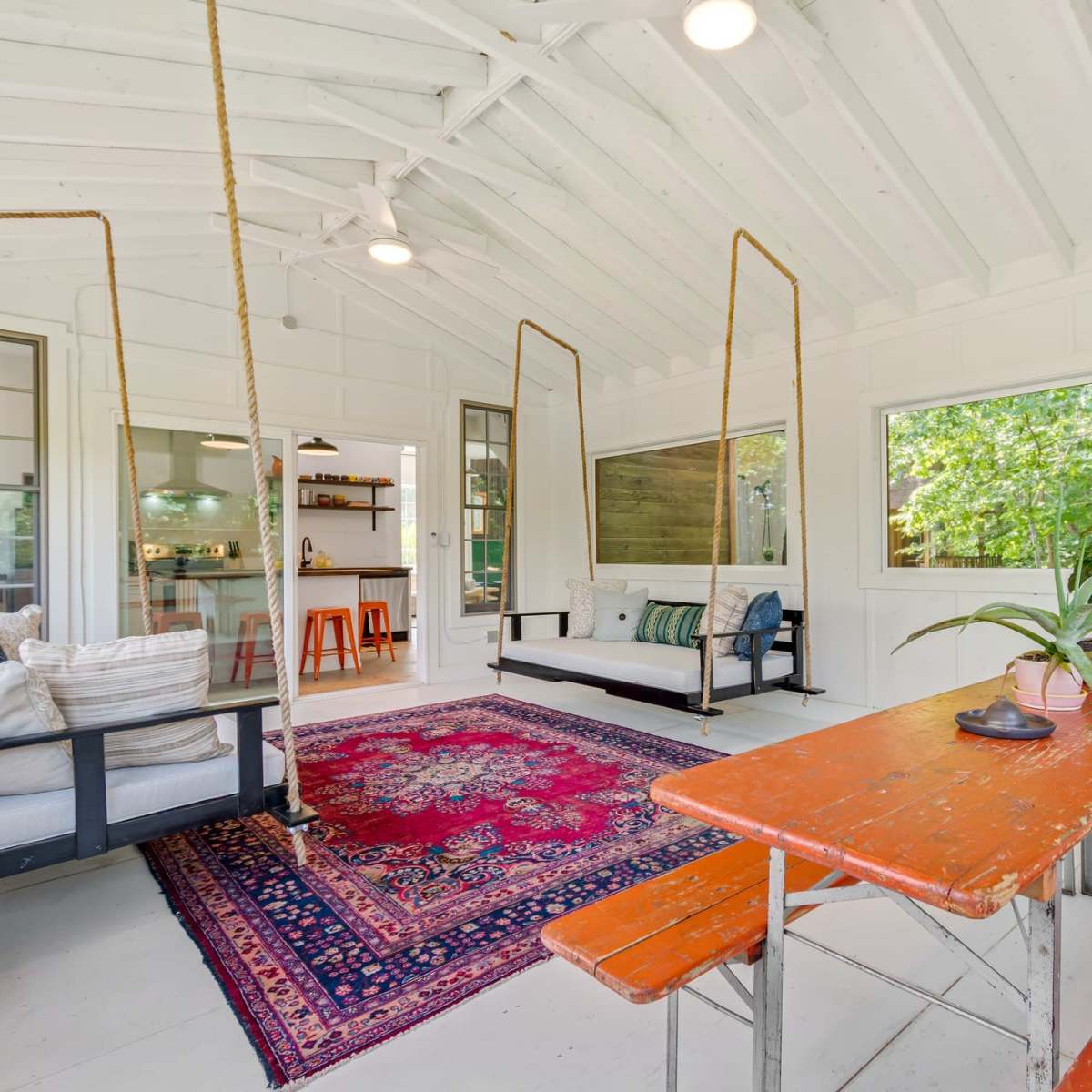 The interior of a bright, open space features a rustic table, hanging swings, and large windows overlooking greenery.