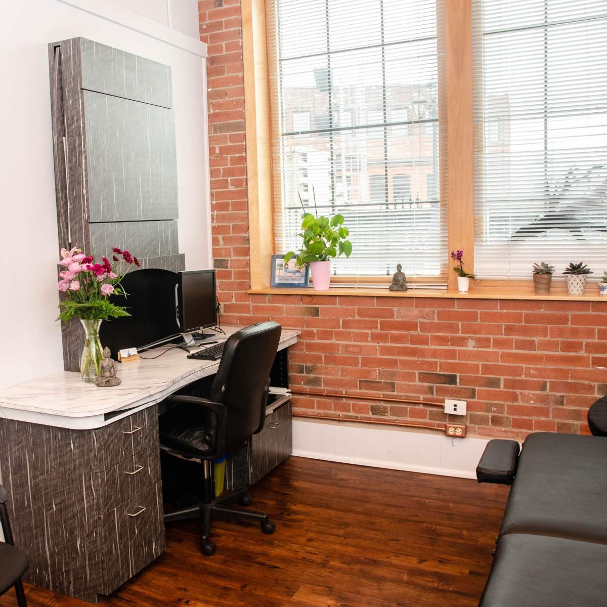A therapy office featuring a desk with a computer, a chair, and a treatment table, all set against a backdrop of exposed brick walls and large windows.