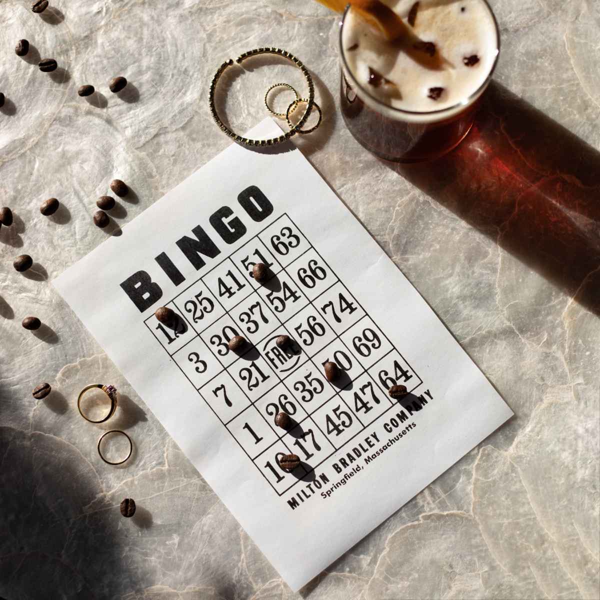 A bingo card is placed on a textured surface alongside coffee beans, a ring, and a glass of iced coffee.
