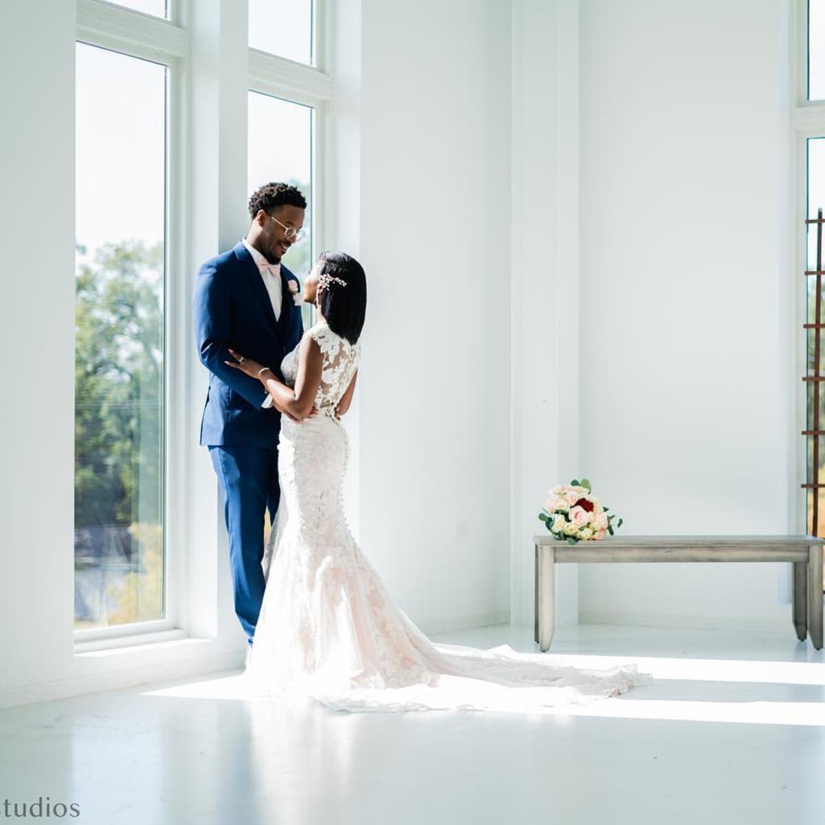 A couple dressed in wedding attire stands together in a bright, modern room with large windows and a simple wooden bench.