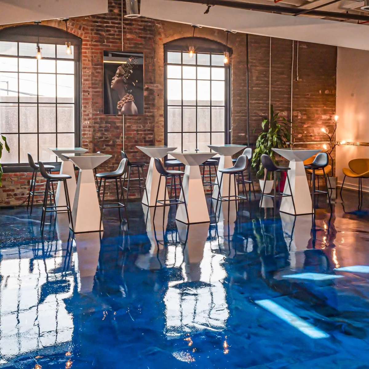 A modern interior space with a glossy blue floor, featuring tall white tables and black and yellow chairs, and large windows allowing natural light to enter.