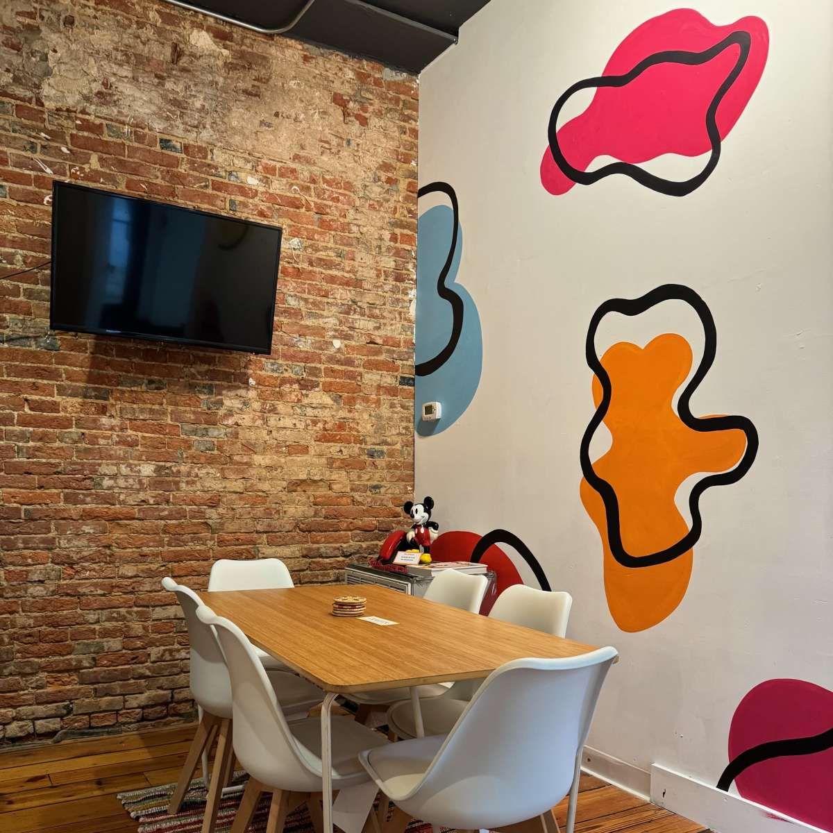 A modern meeting room with a wooden table surrounded by white chairs, a brick wall, and colorful abstract shapes painted on the wall.