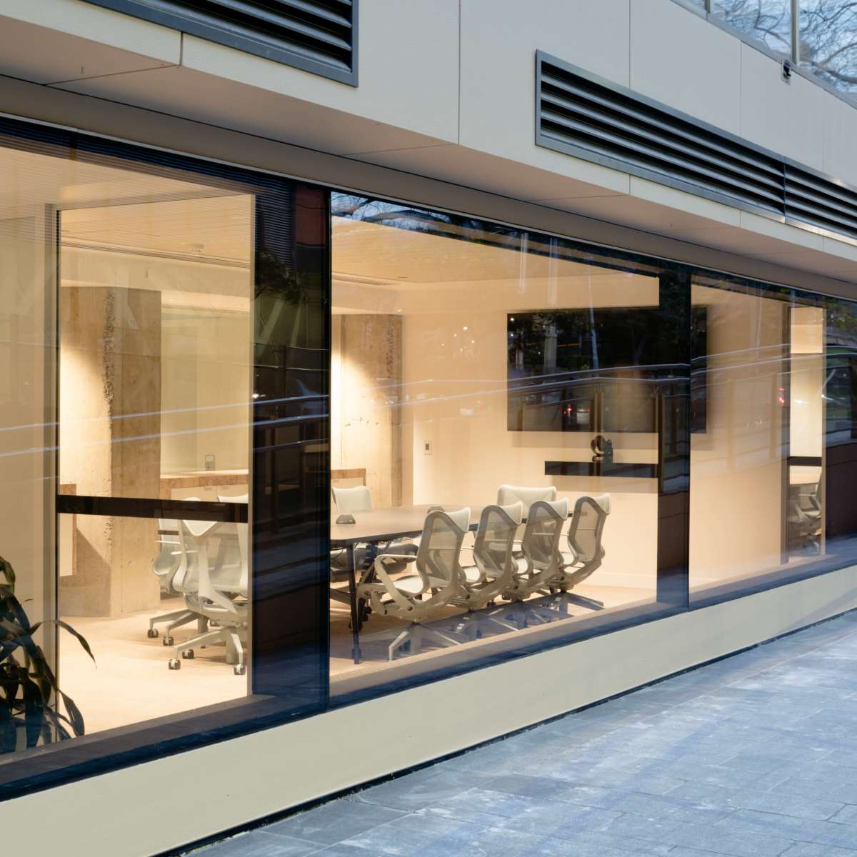 A modern office conference room with glass walls, a long table, and ergonomic chairs, visible from the exterior.