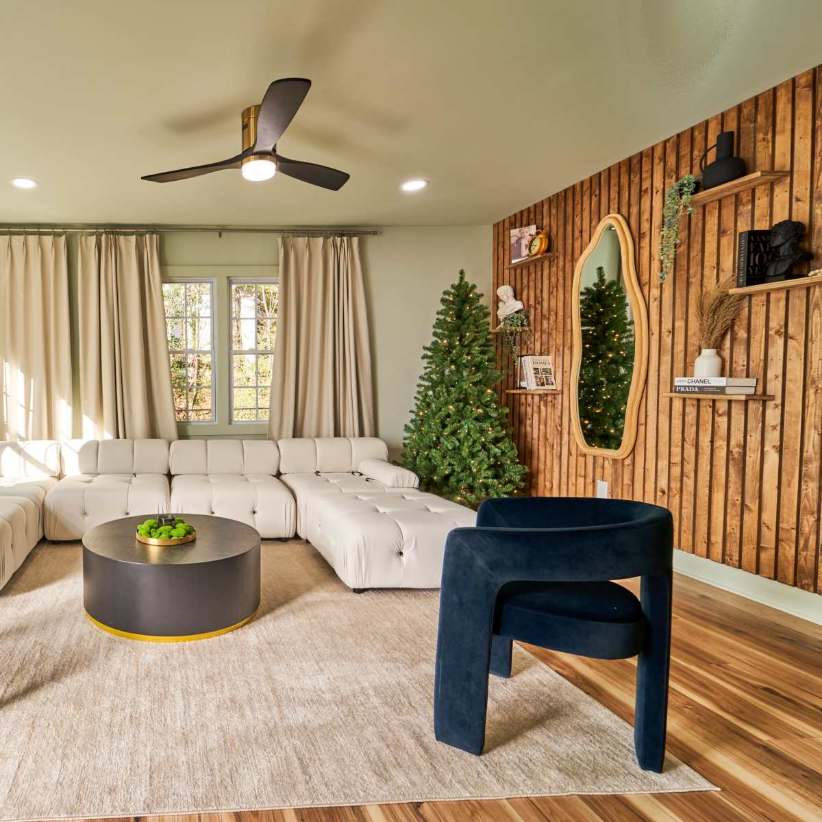 A modern living room features a white sectional sofa, two blue chairs, a round coffee table, and decor against a wooden accent wall.