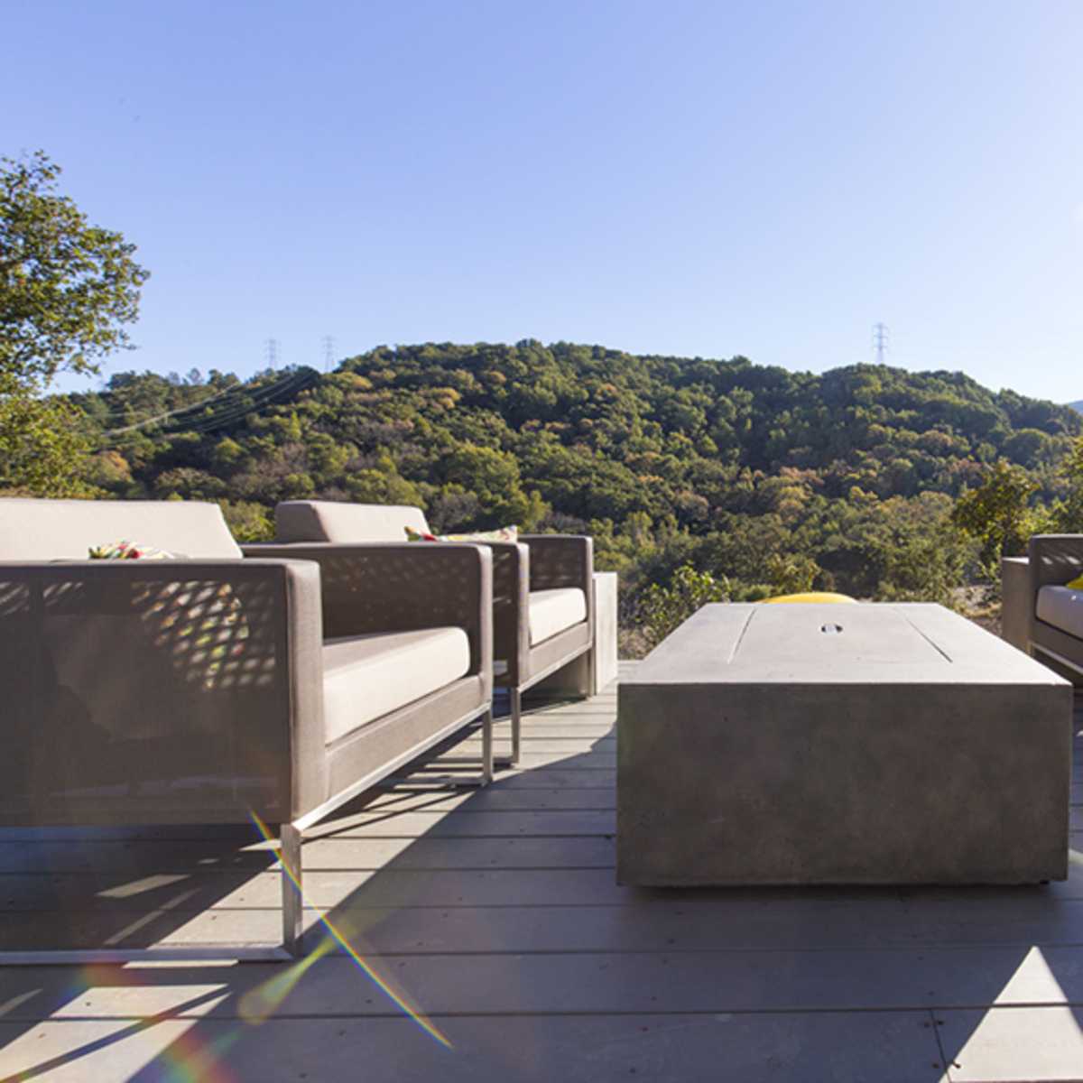 A modern outdoor seating area features two sofas and a rectangular coffee table, surrounded by lush green hills and clear blue skies.