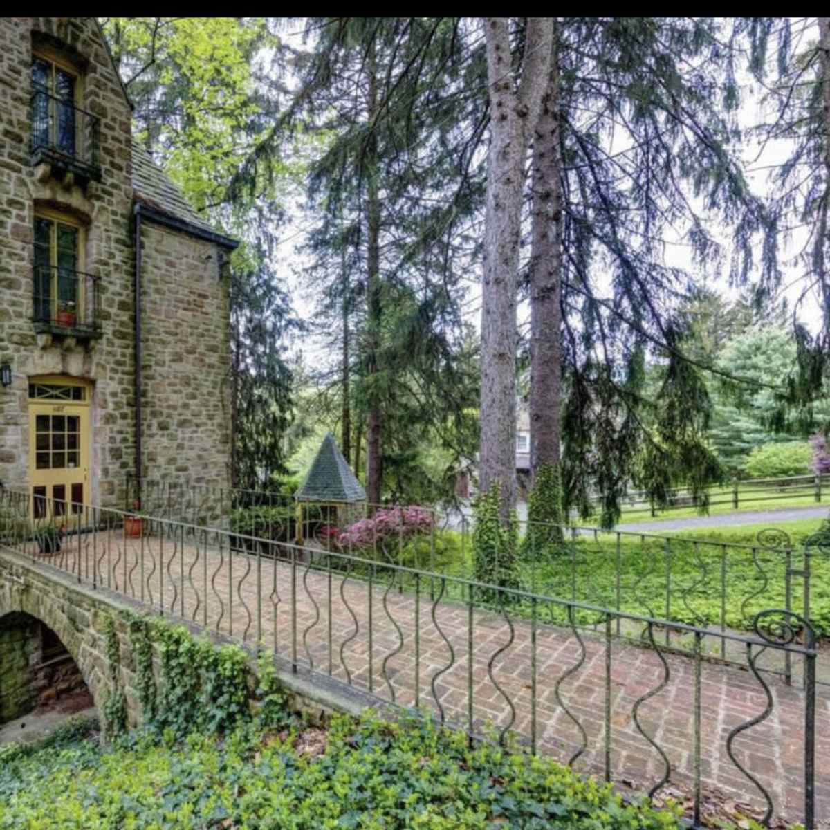 A stone house with an arched bridge leads to its entrance, surrounded by tall trees and green landscaping.