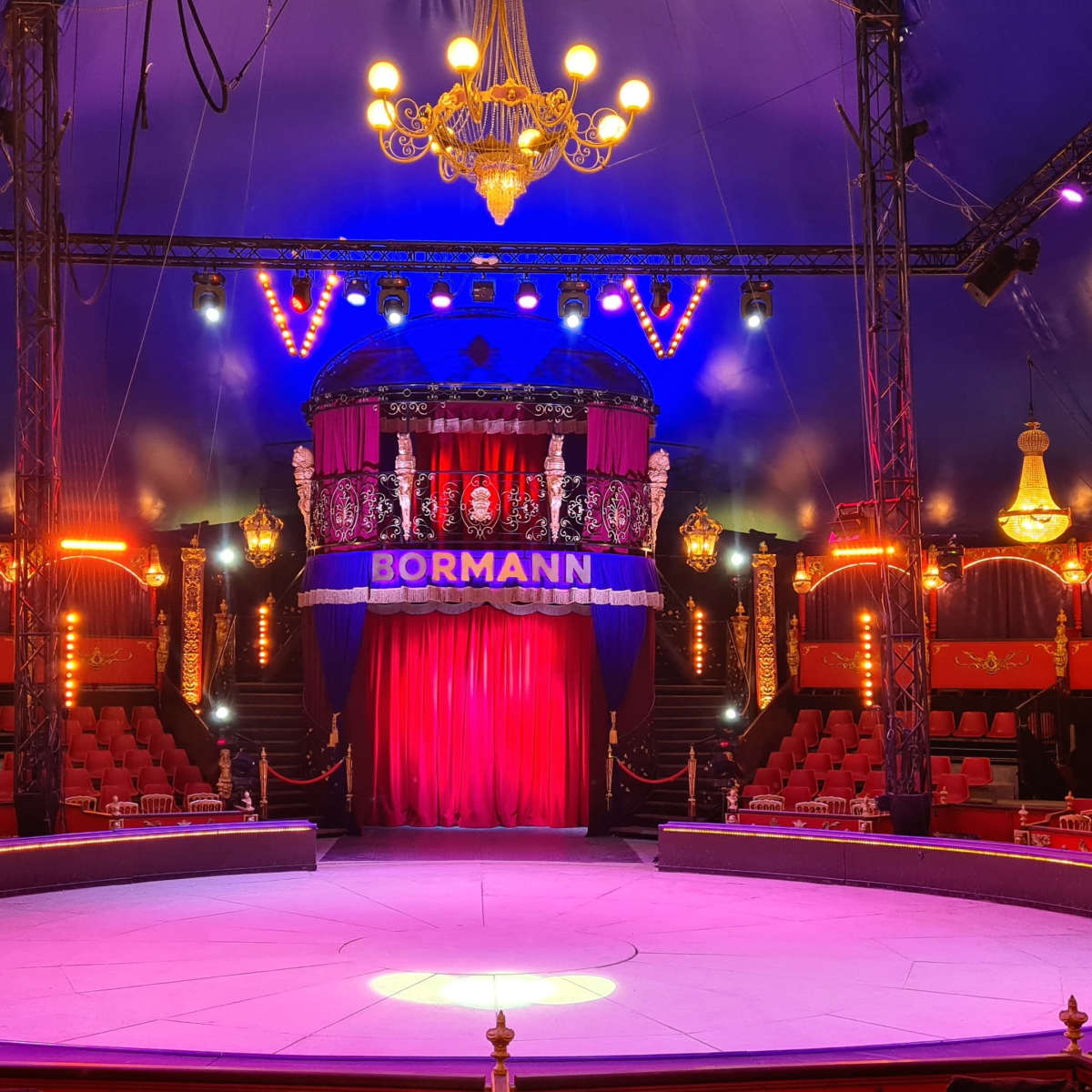 A brightly lit circus tent with a central performance ring surrounded by seats and decorative stage elements.