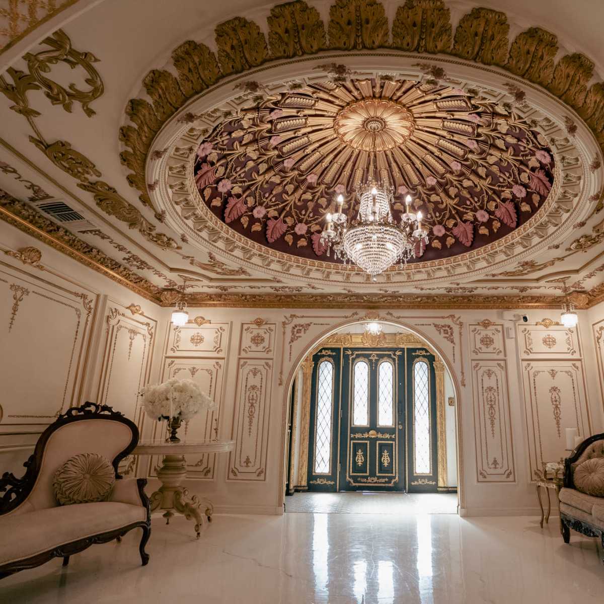 An ornate foyer with a chandelier, detailed ceiling, and ornate furniture, leading to a set of double doors.