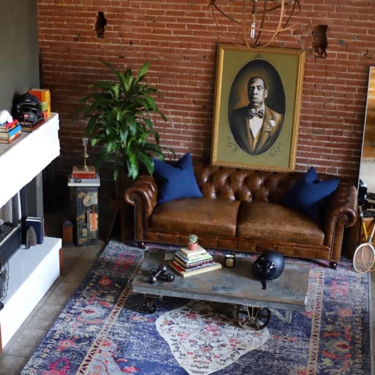 A living room with a brown leather sofa, a coffee table adorned with books, and a large portrait hung on a brick wall.