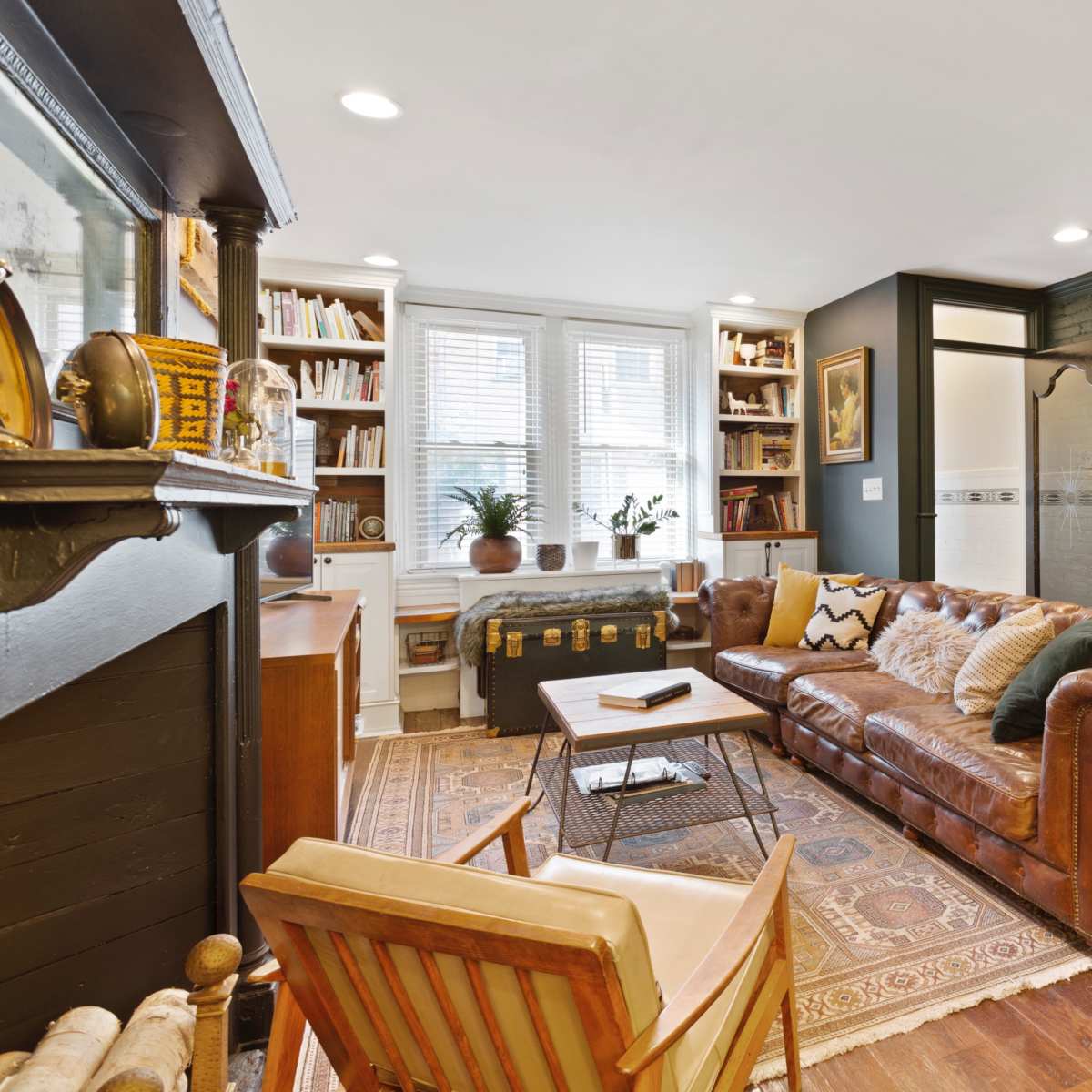 A cozy living room featuring a brown leather sofa, a wooden armchair, a bookshelf, and decorative items against a dark green wall.
