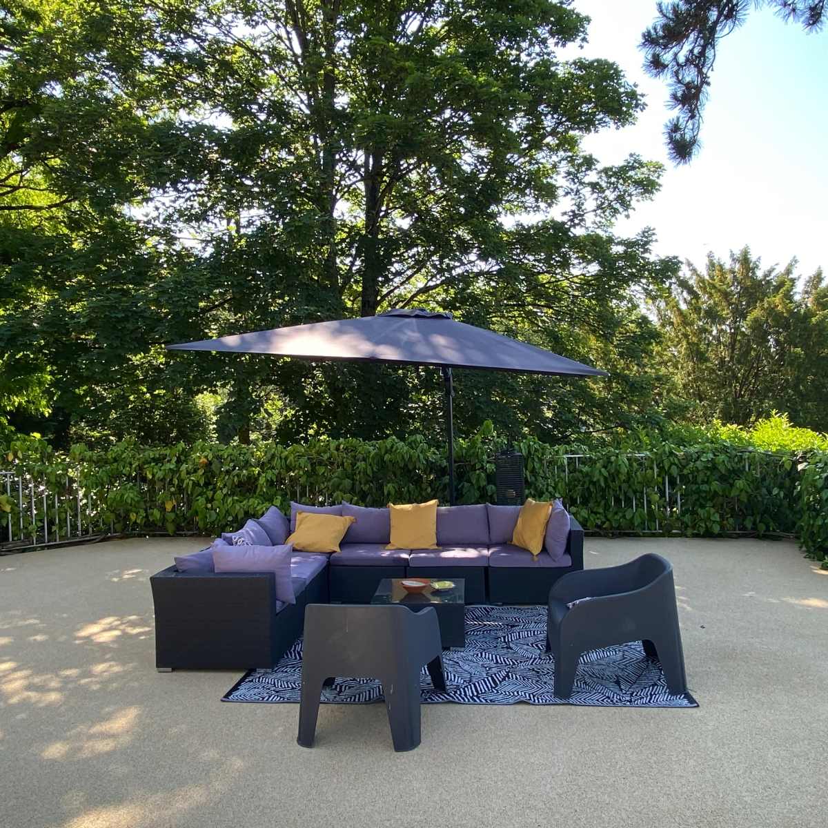 A patio area is furnished with a sectional sofa, two chairs, a coffee table, and a large umbrella, surrounded by greenery.