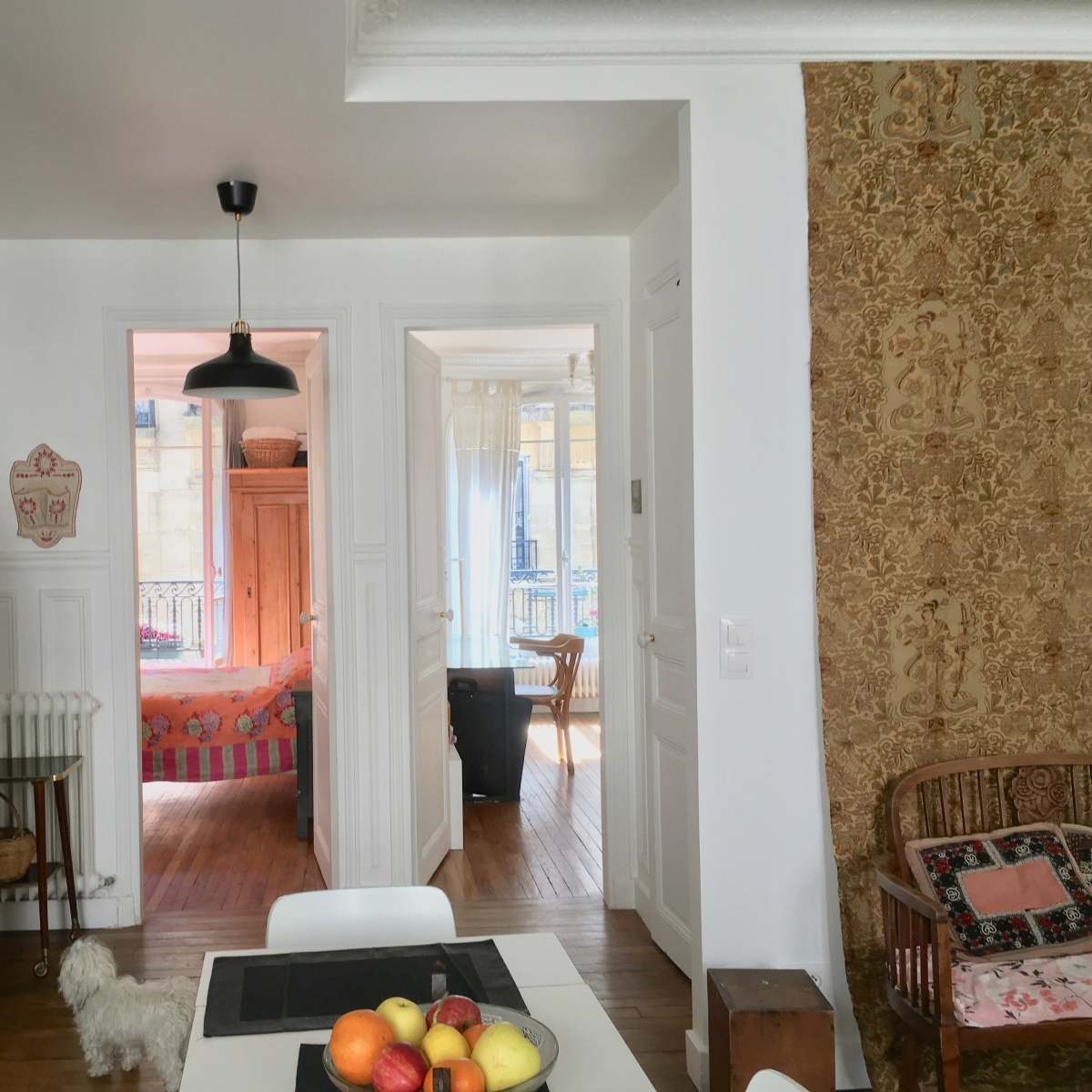 A bright kitchen with a table featuring a bowl of fruit, leading to a hallway that connects to another room with a visible bed.