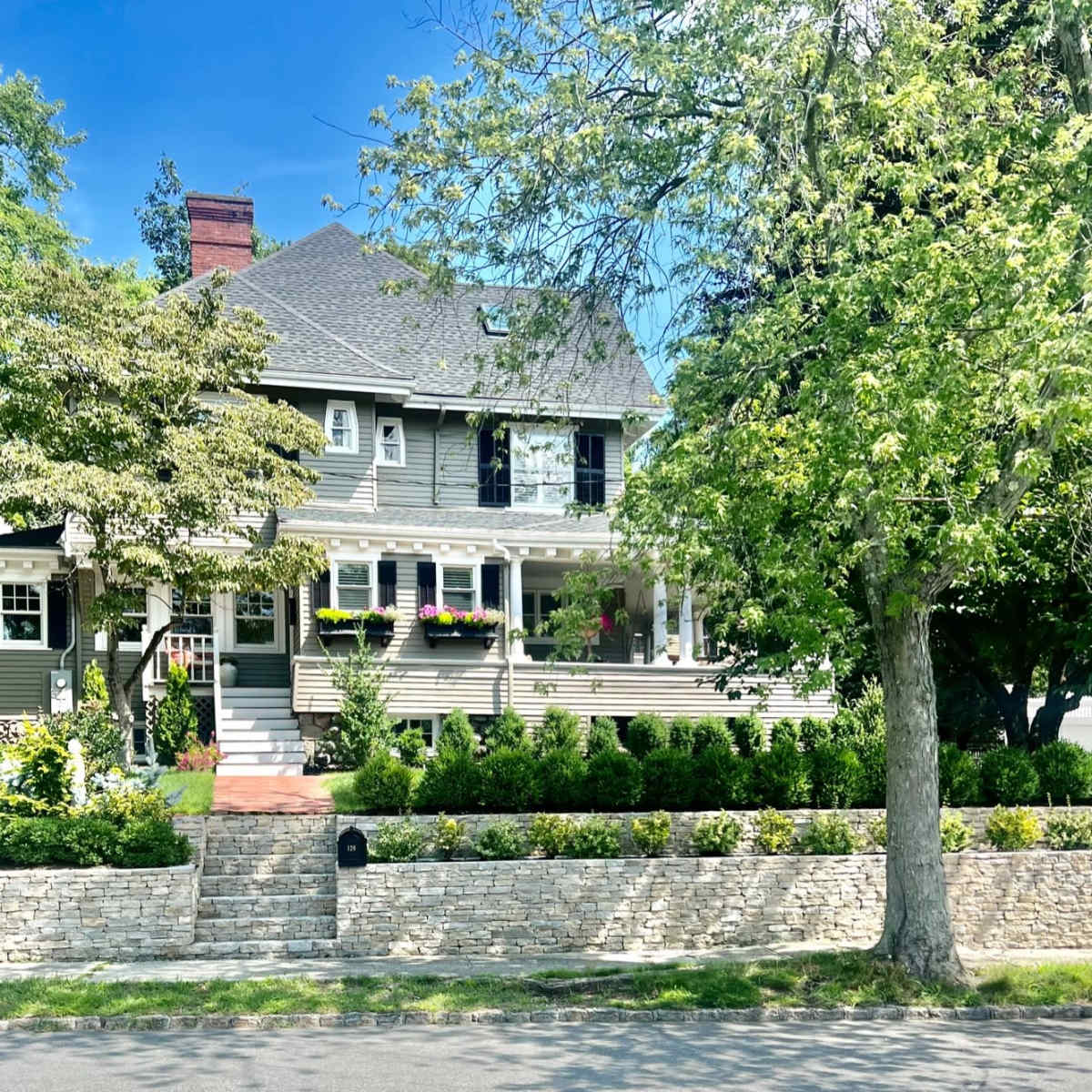 A large, two-story gray house with a front porch and well-manicured gardens is located at the end of a quiet street.