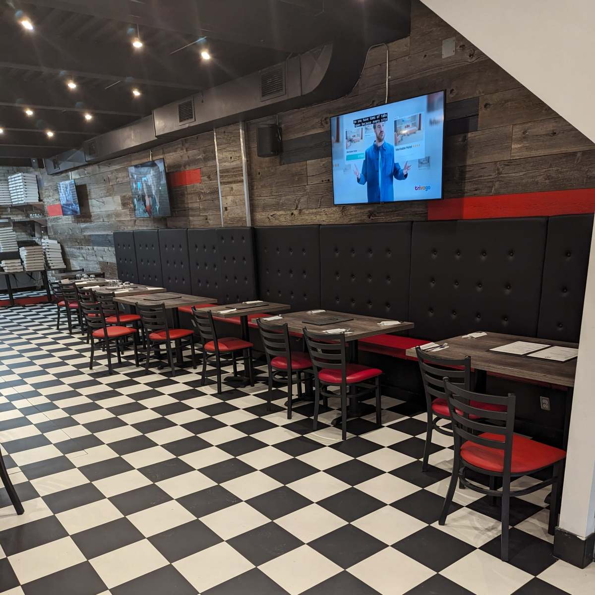 An empty dining area with black and white checkered flooring, red-accented booths, and two televisions mounted on the wall.