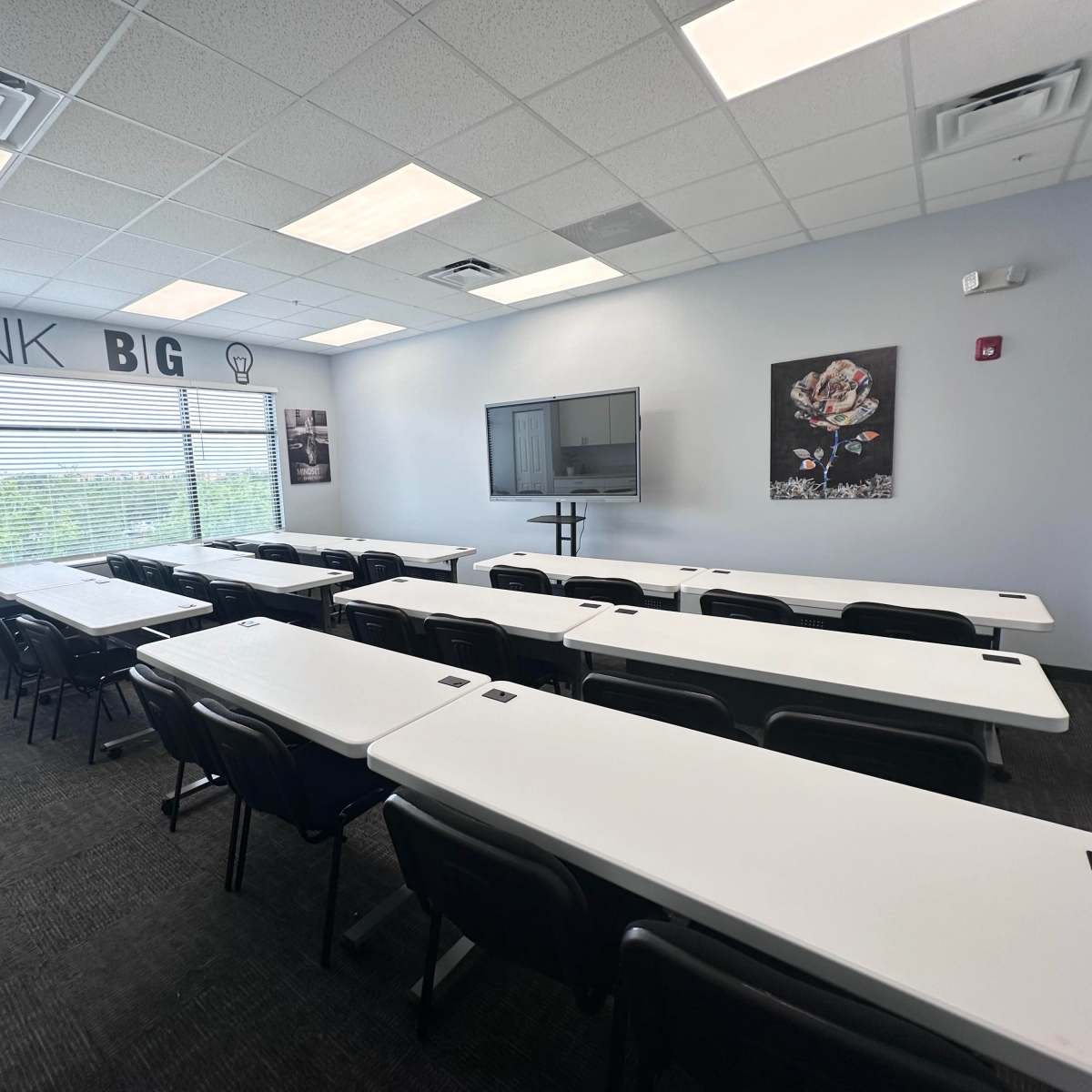 A modern classroom is set up with multiple tables and chairs arranged in rows, featuring large windows and a wall with motivational text.