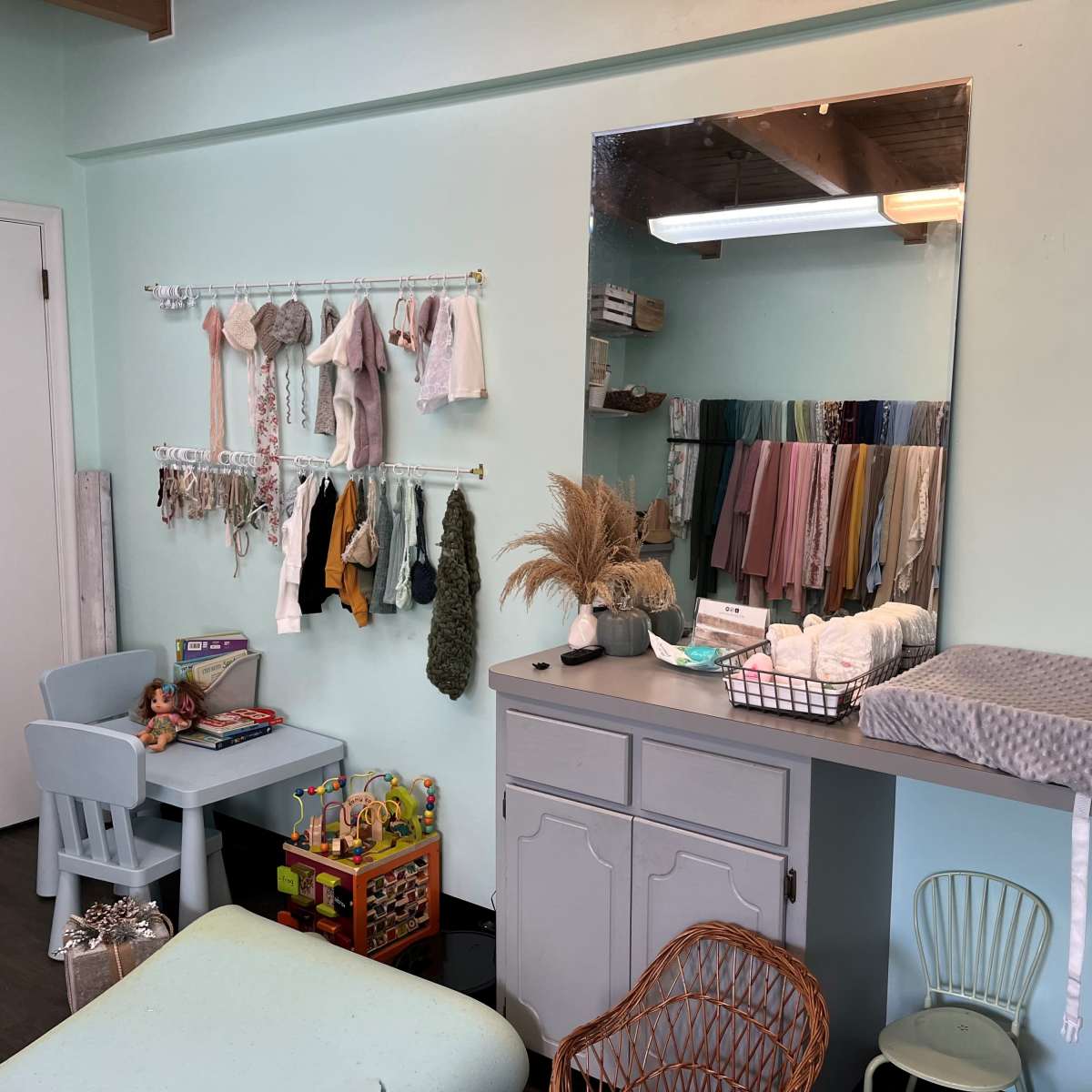 A light blue room with a changing table, a mirror, various clothes hanging on a rack, and children's toys in a corner.