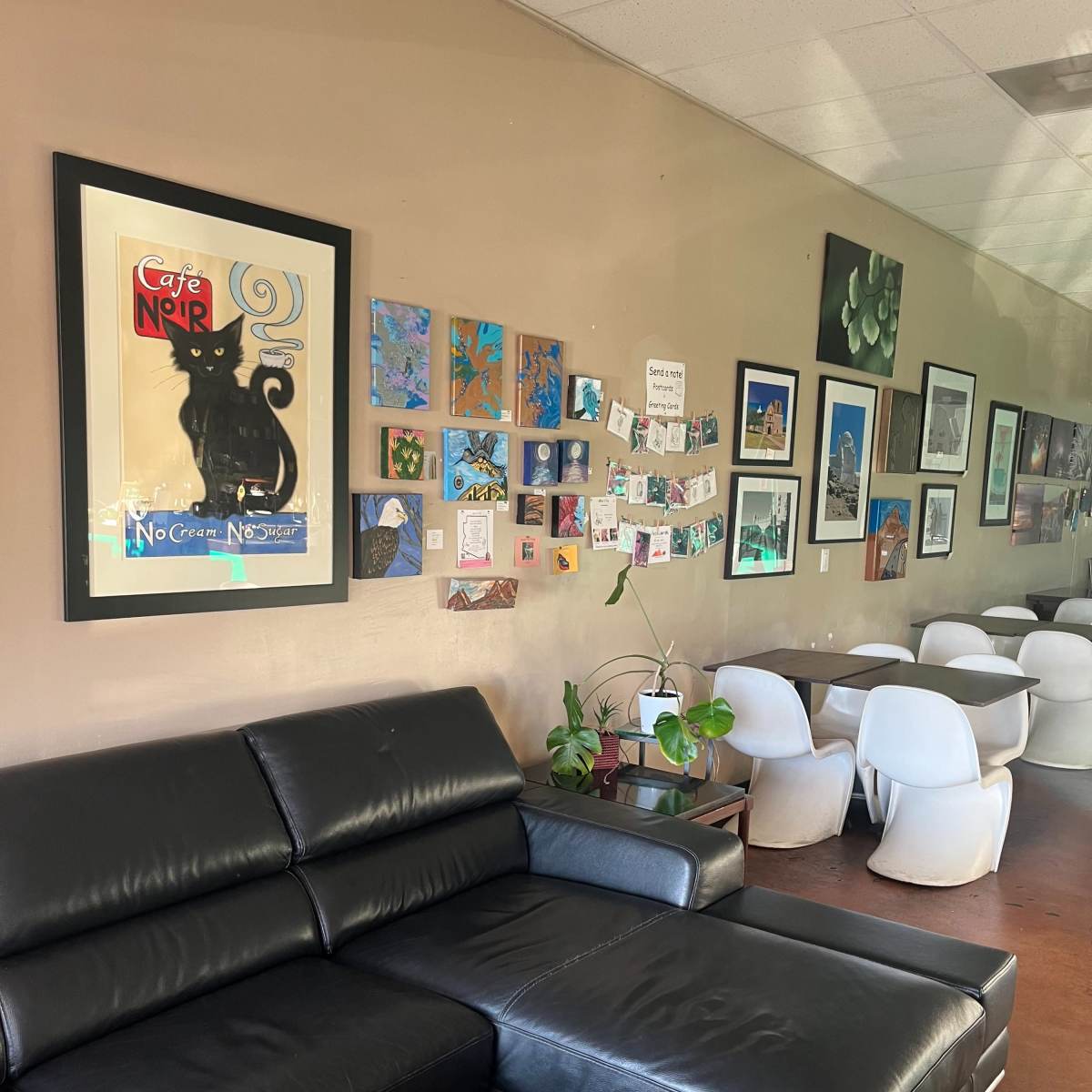 A cafe interior with a black leather sofa, a wall decorated with various framed artworks, and several tables and chairs in the background.