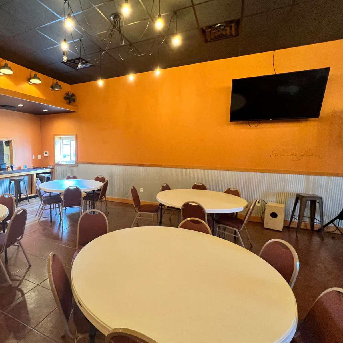 A dining area with round tables and chairs, orange walls, and a bar counter in the background.