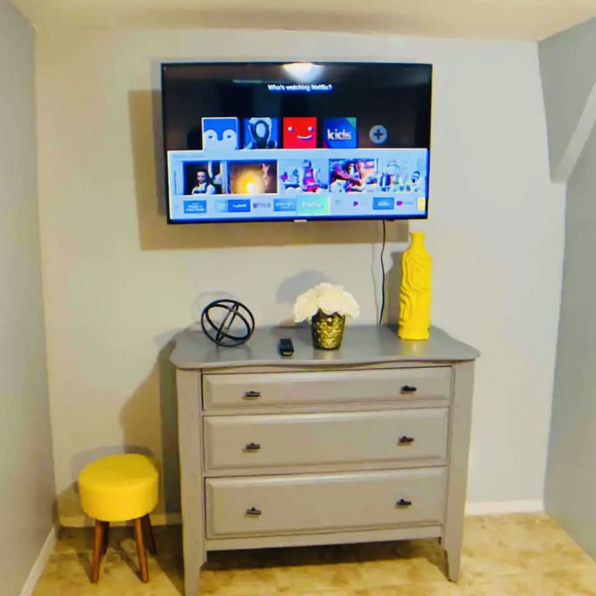 A wall-mounted TV displays various menu options above a gray dresser with a yellow decorative vase and a small yellow stool nearby.