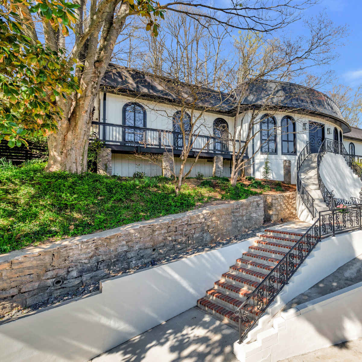 A large, modern house with a curved roof is set on a hillside, featuring a stone staircase leading to the front entrance.