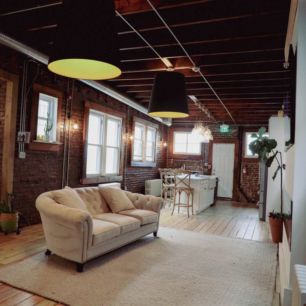 A spacious room with exposed brick walls, featuring a cream-colored couch on a light rug, with large windows and a kitchen area visible in the background.