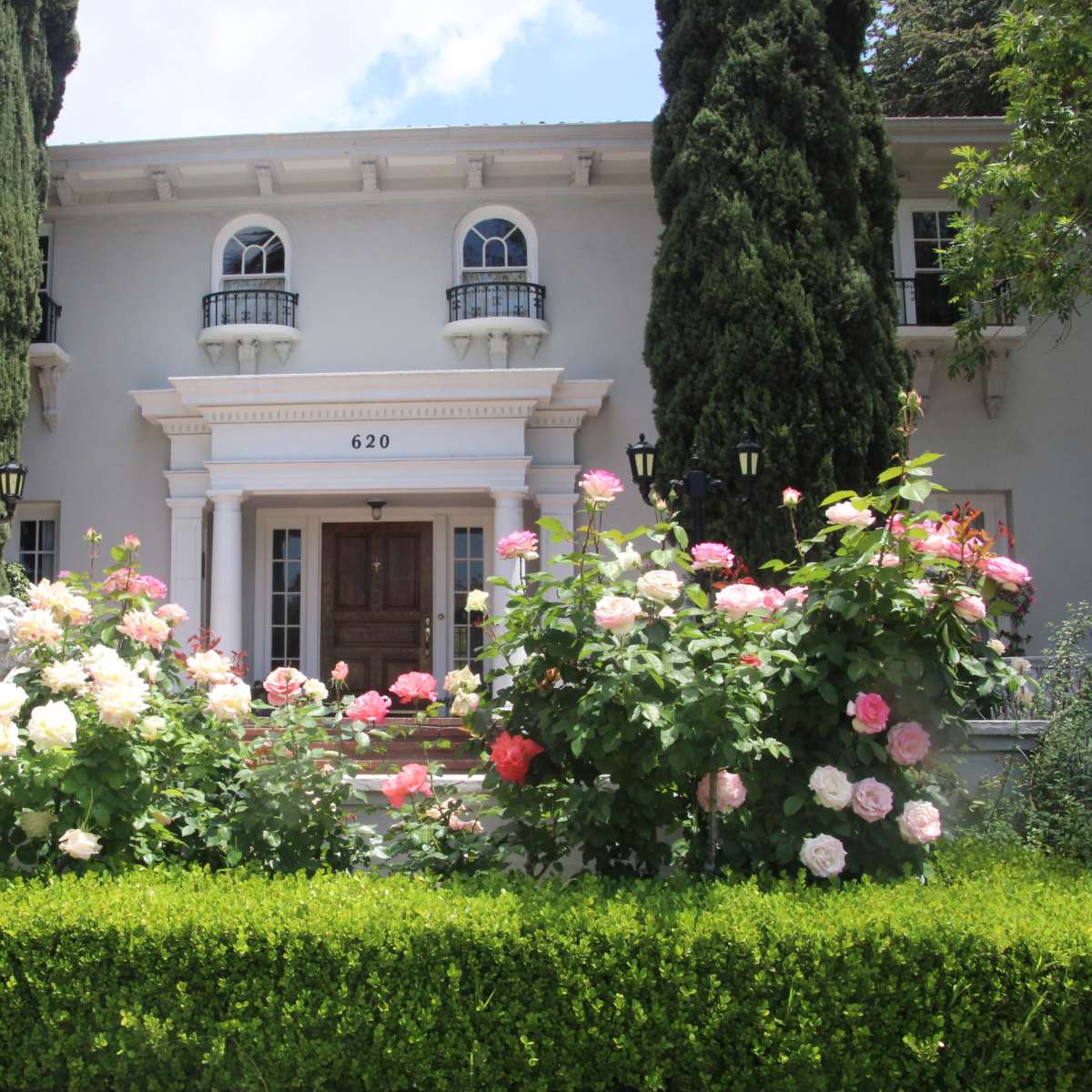 A large, two-story house with a columned entrance and surrounded by tall cypress trees and vibrant rose bushes.
