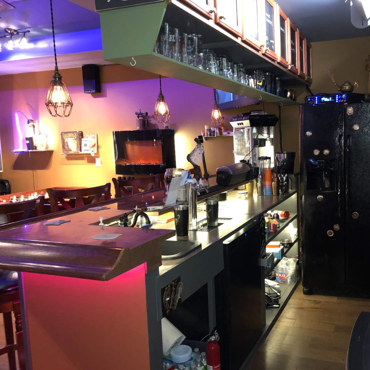 A cozy bar area with a counter, shelves lined with bottles, and a black refrigerator in a warmly lit room.