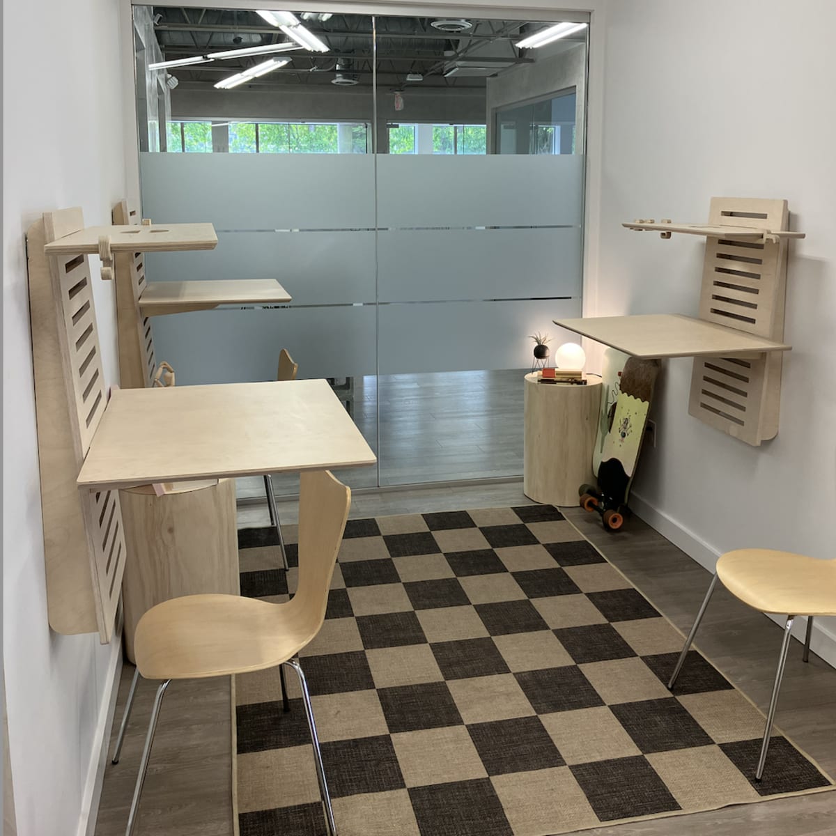 A minimalist workspace with two wooden desks attached to the walls, each accompanied by a chair, and a checkered rug on the floor.