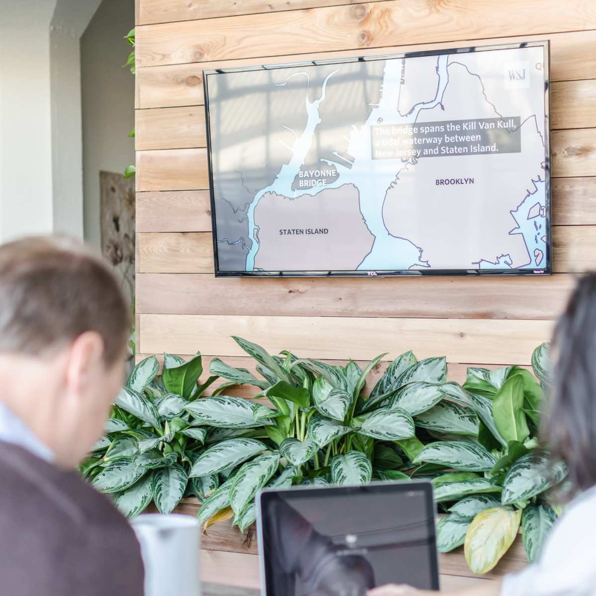 Two individuals sit at a wooden table with laptops and cups of coffee, while a map is displayed on a wall-mounted screen behind them.