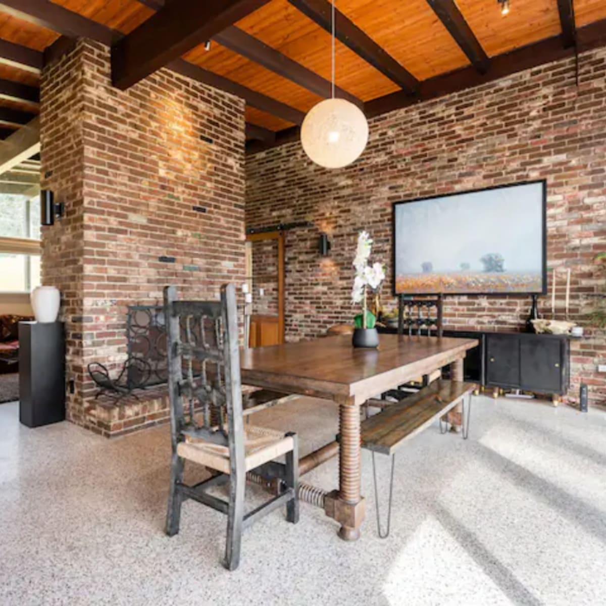 A modern living space with exposed brick walls, a wooden dining table, and large windows allowing natural light to enter.