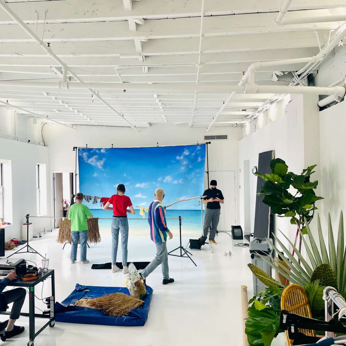 A film studio setup is shown with a blue backdrop depicting a beach scene, while several people and equipment are arranged for a photoshoot.