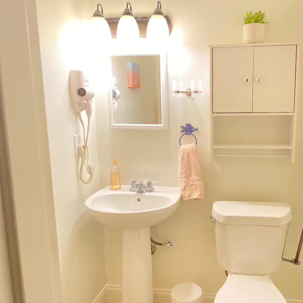 A small bathroom featuring a sink with a mirror above it, a toilet, a wall-mounted shelf, and a hairdryer attached to the wall.