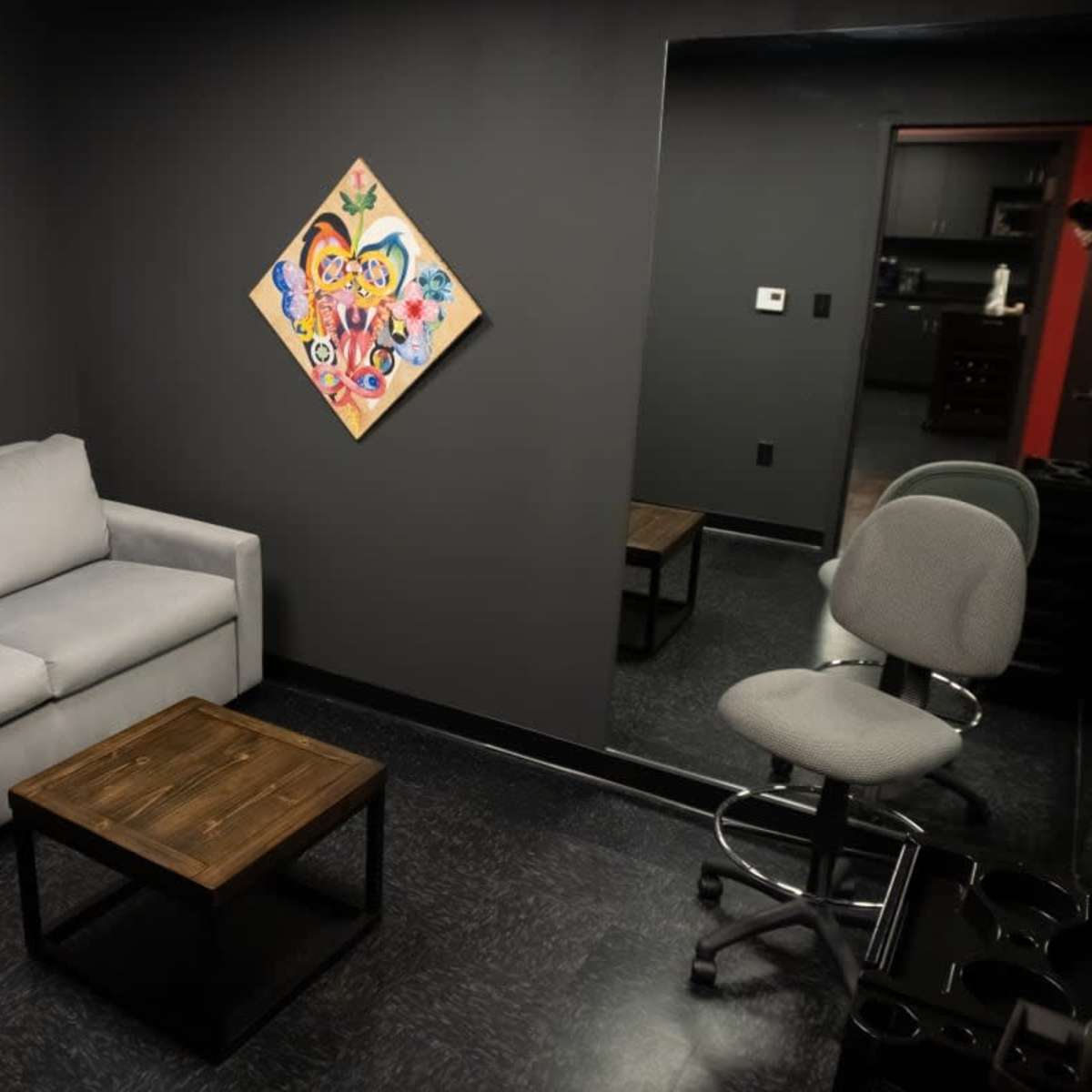 A small, dark room with a gray sofa, a wooden table, a swivel chair, and a colorful painting on the wall.