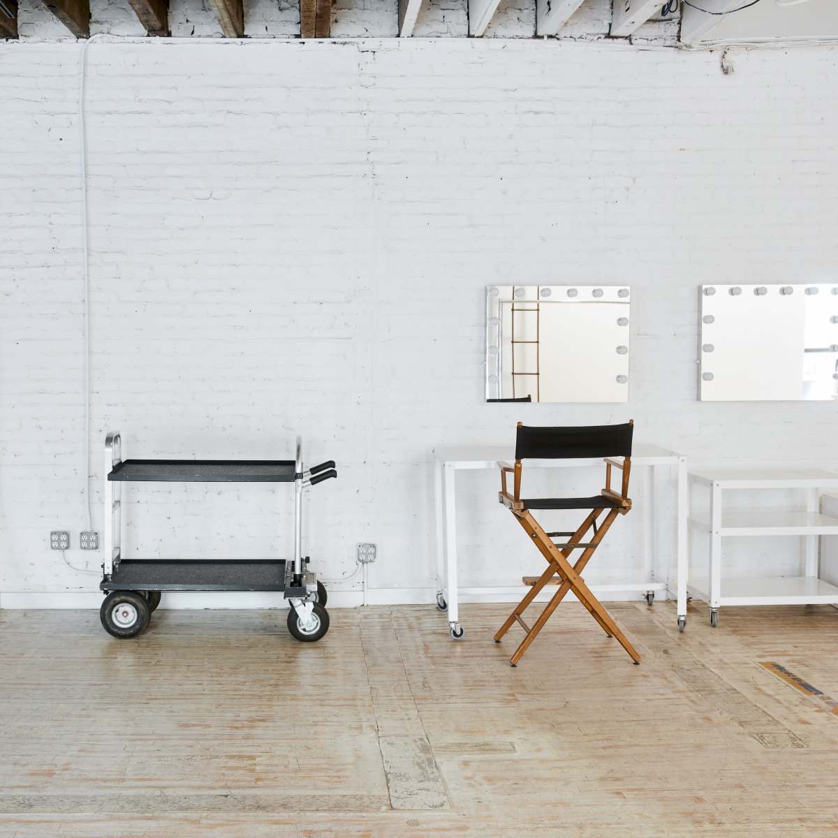 A minimalist studio space with a white backdrop, a cart on wheels, a director's chair, and two illuminated mirrors on a table.