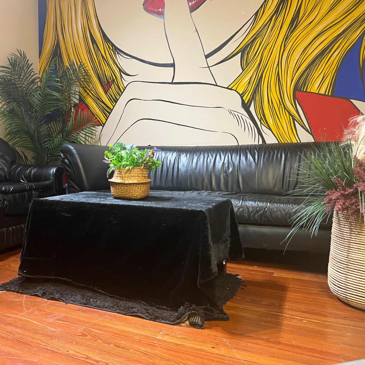 A room with a black leather couch and a black table covered with a velvet cloth, surrounded by decorative plants and a vibrant mural on the wall.