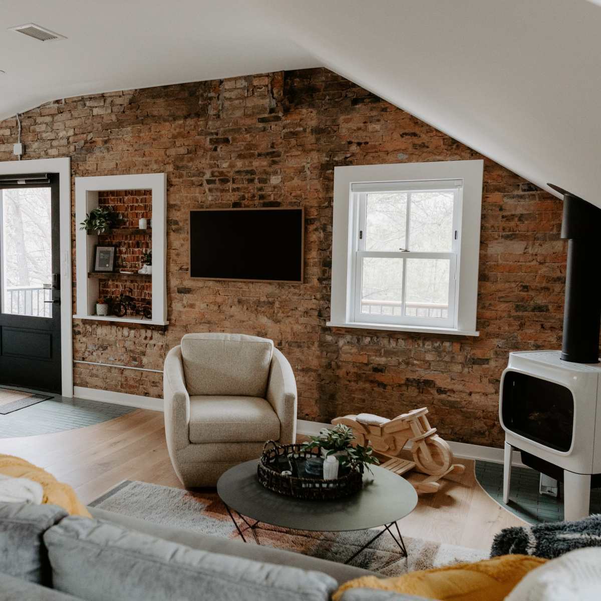 A cozy living room features a brick wall, modern furnishings, and a large window with natural light.