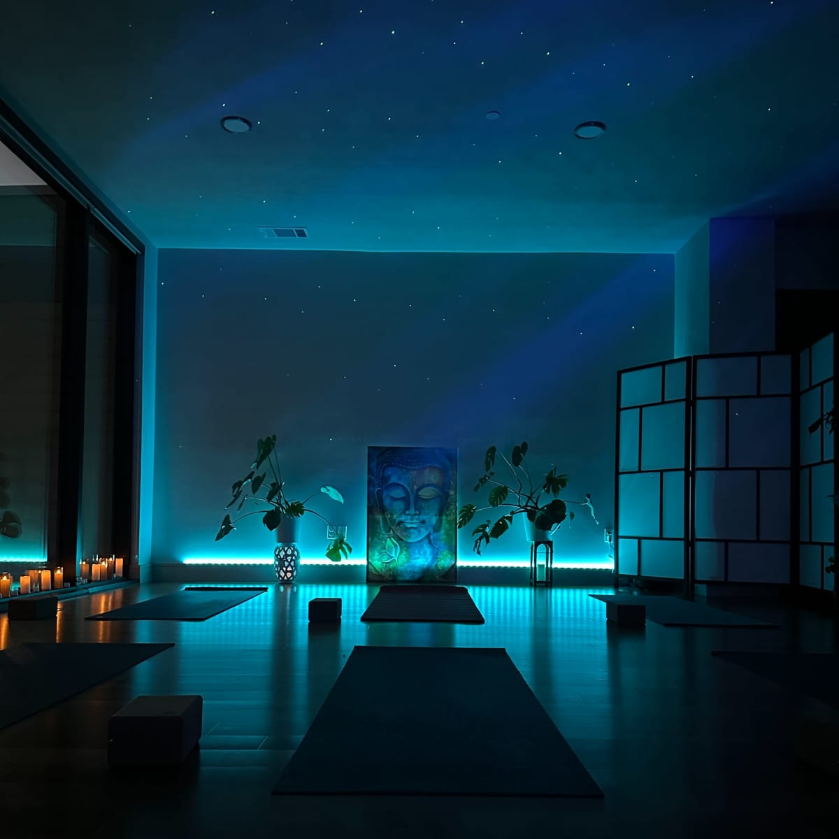 A serene yoga studio featuring a central Buddha painting, illuminated by soft blue lighting, surrounded by plants and candles.