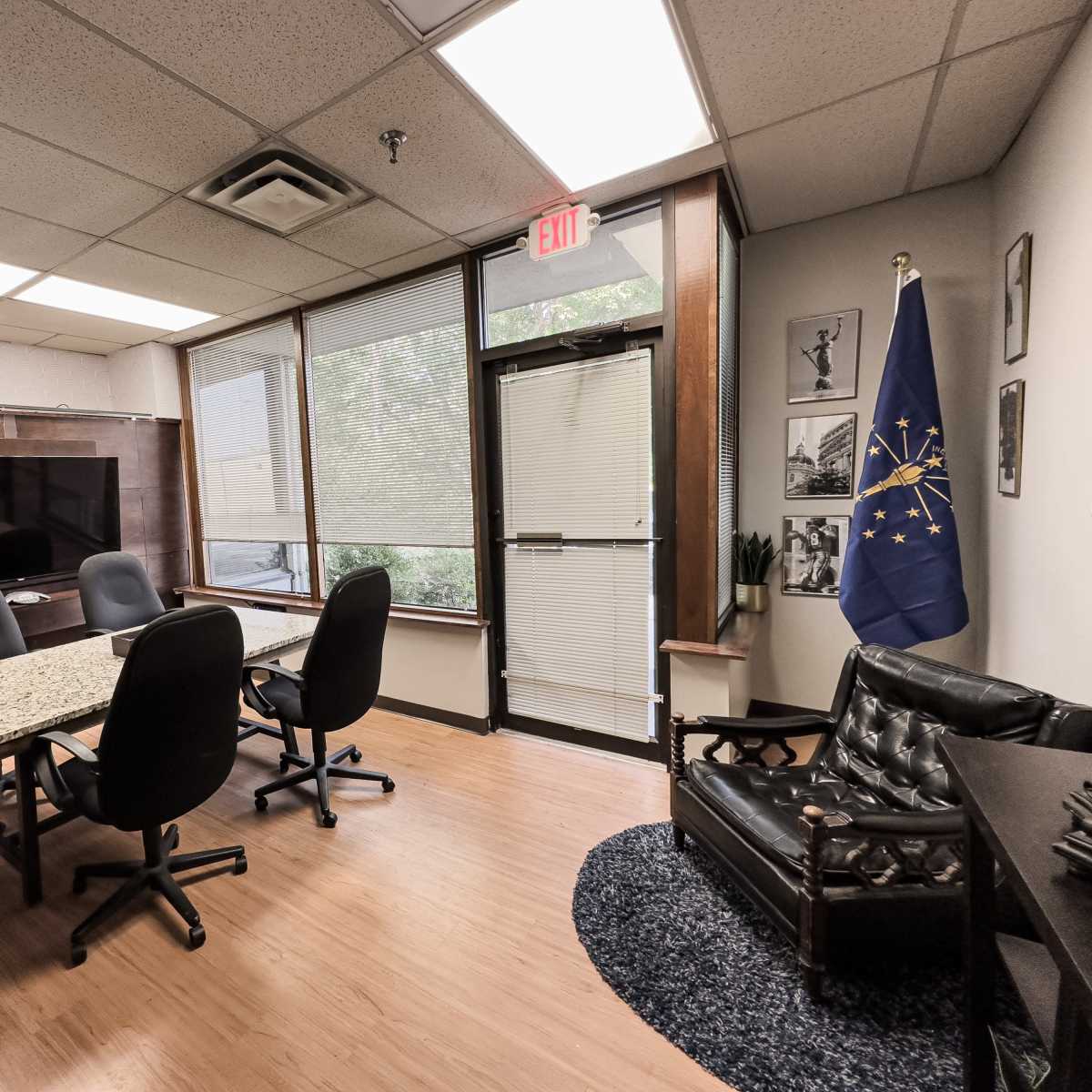 A conference room featuring a large table surrounded by black chairs, a television mounted on a wooden wall, a door leading outside, and a flag displayed in the corner.