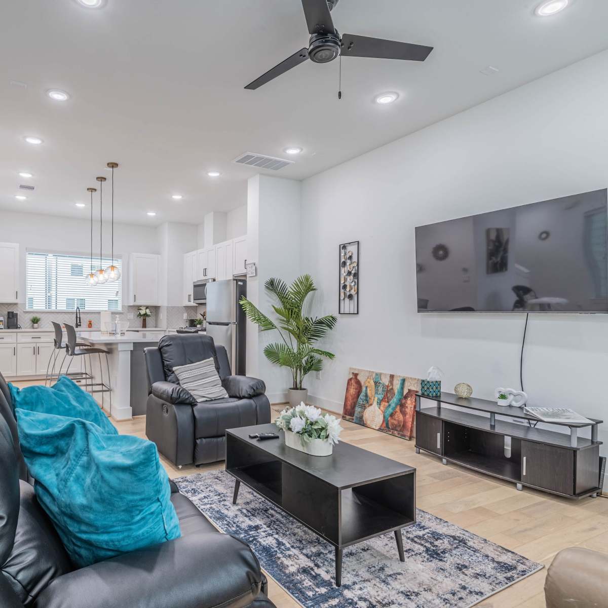 A modern living room with black recliners, a television mounted on the wall, a coffee table, and a kitchen area visible in the background.