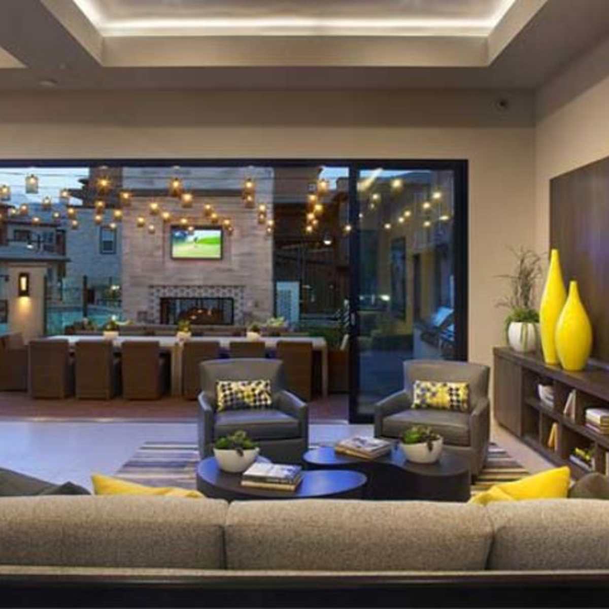 A modern living room features a large sectional sofa, a coffee table, and a view of an outdoor dining area illuminated by string lights.