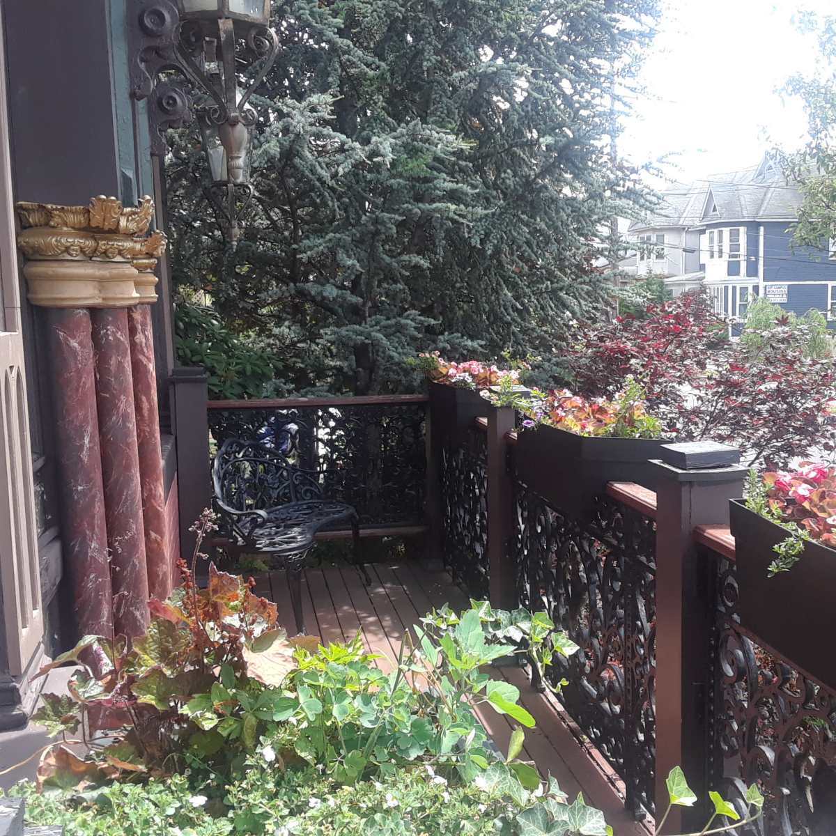 A balcony with decorative railings and planters filled with greenery overlooks a garden and nearby houses.