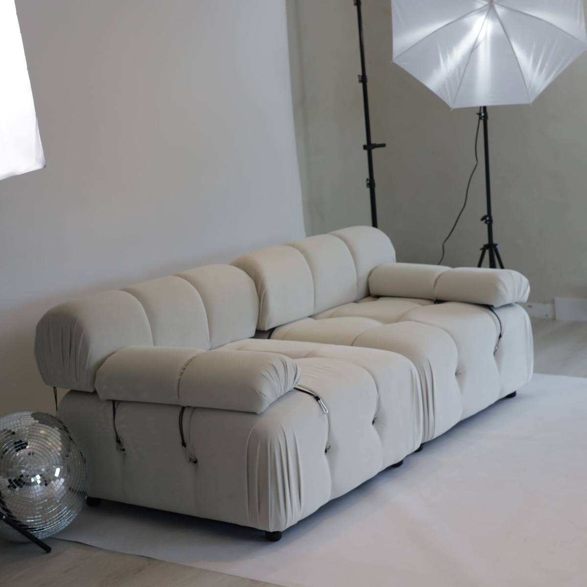 A beige sectional sofa is positioned in a minimalistic studio setting, accompanied by two softbox lights and a mirrored disco ball.