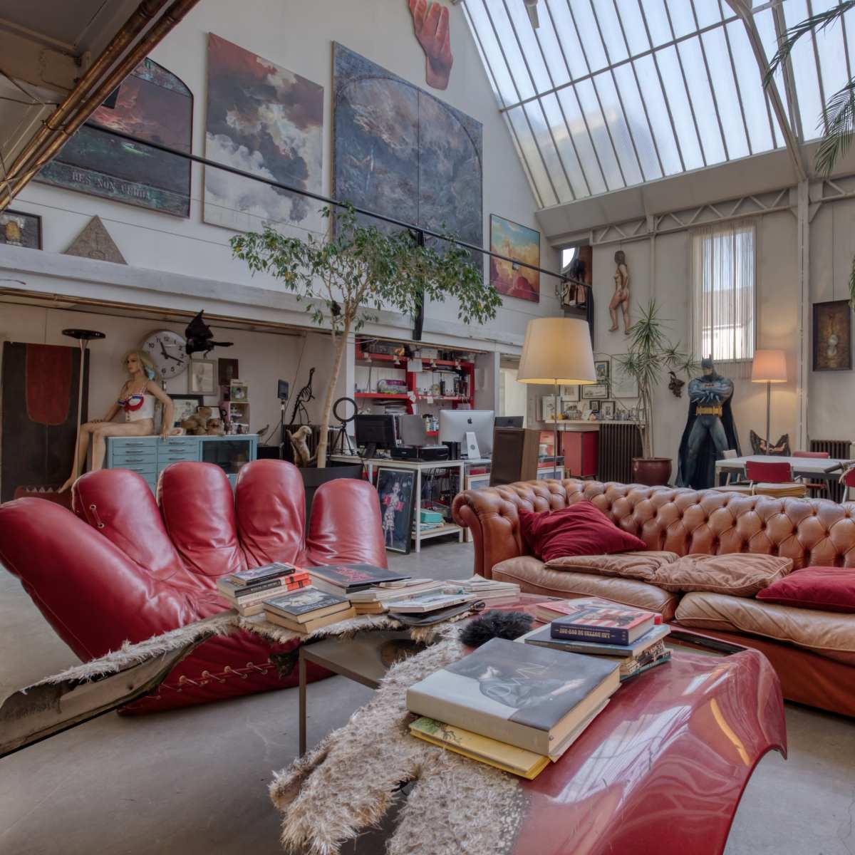 A spacious, eclectic living room featuring a red chesterfield sofa, a glass coffee table piled with books, and various artwork on the walls.