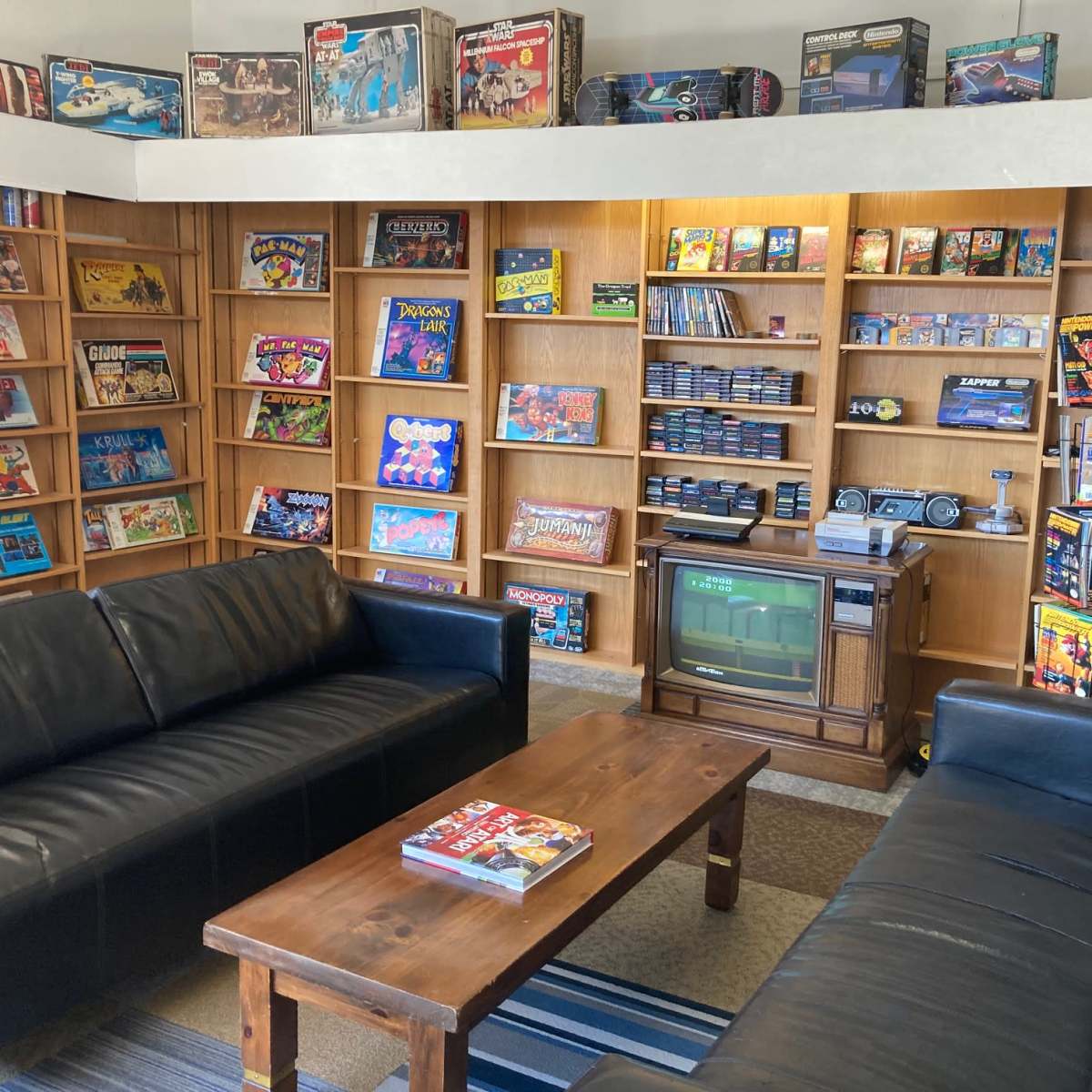 A cozy game room with two black sofas facing a wooden coffee table, surrounded by shelves filled with board games and video game consoles.
