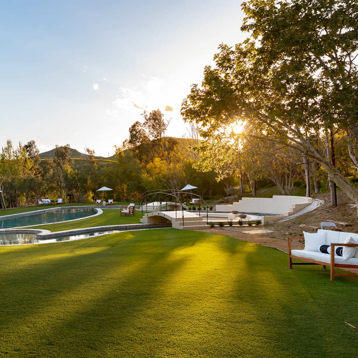 A landscaped outdoor area featuring a swimming pool, lounge chairs, and trees, with sunlight filtering through the foliage.