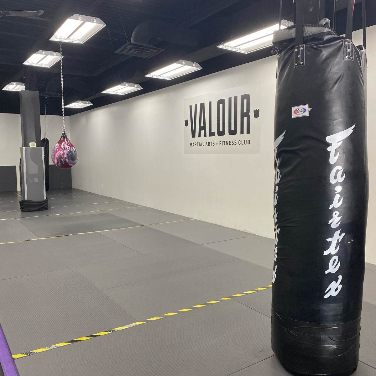 An empty martial arts training room featuring a punching bag, a speed bag hanging from the ceiling, and mats on the floor with a wall displaying the name "Valour Martial Arts & Fitness Club."