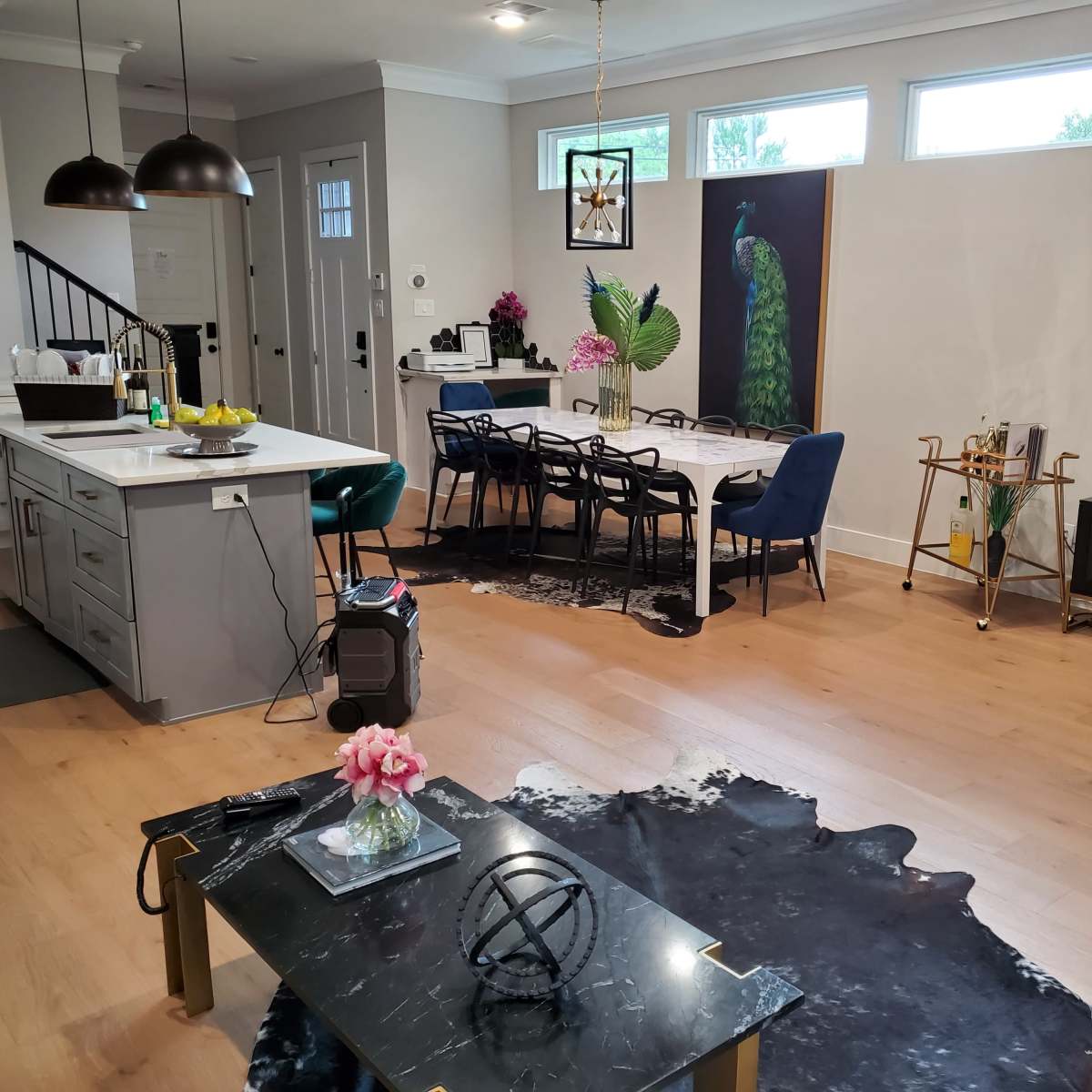 A modern living space featuring a kitchen area, a dining table with chairs, a decorative peacock painting, and a television displaying content on a wooden floor.