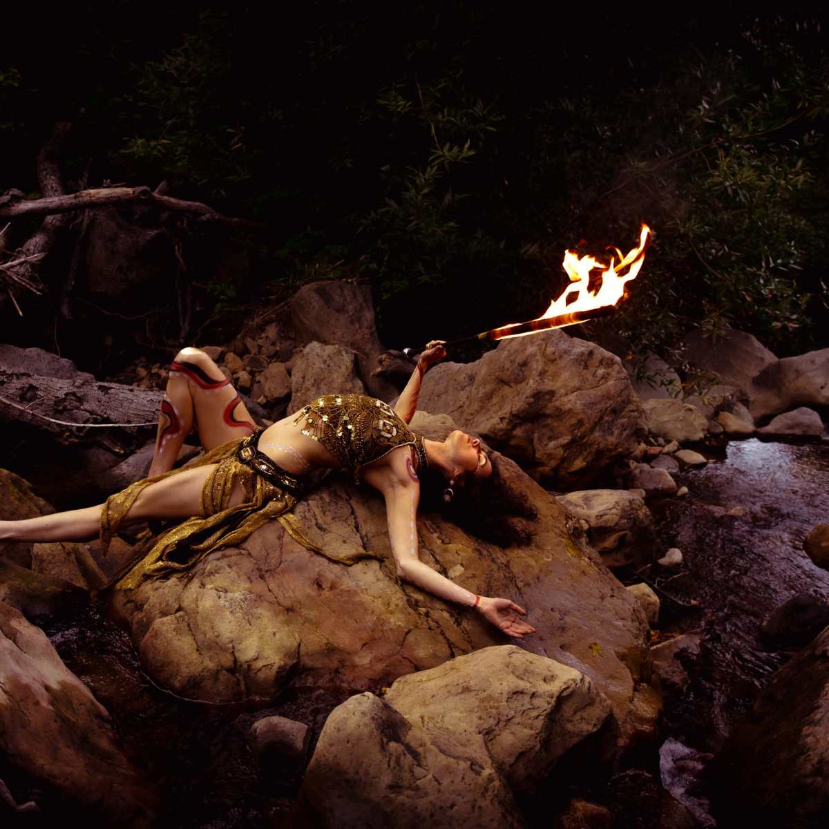 A person lies on a rock near a stream, holding a flaming stick while surrounded by trees and boulders.
