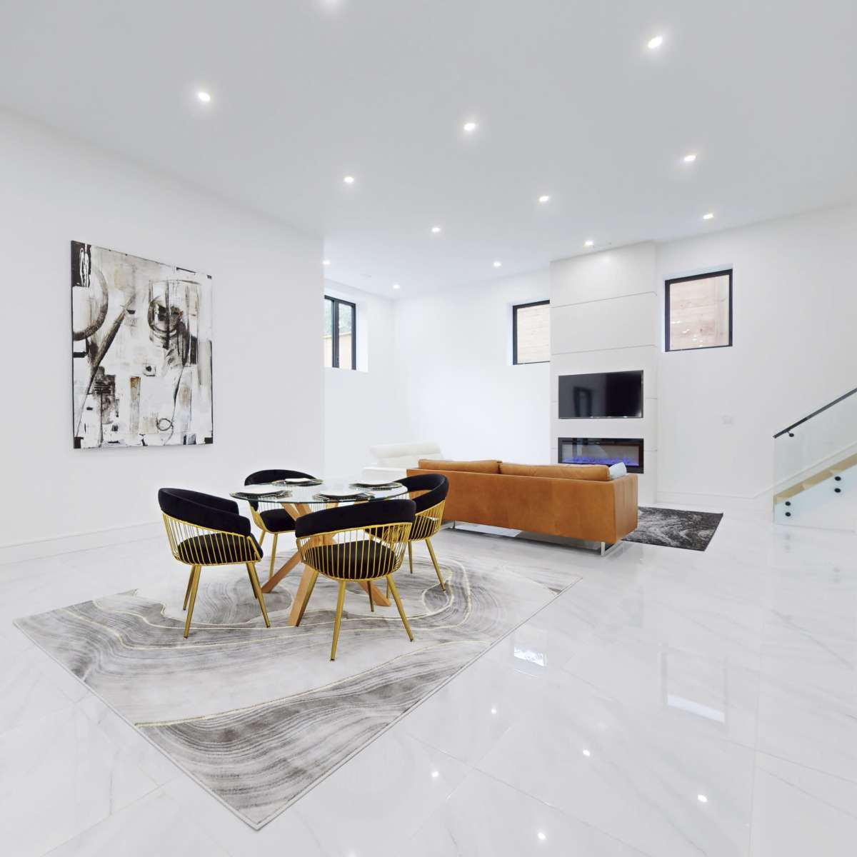 A spacious, modern living area features a round dining table surrounded by black and gold chairs, a large artwork on the wall, and a tan sofa.